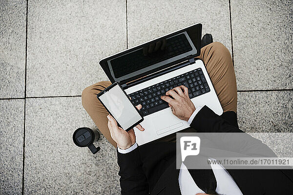 Businessman with mobile phone using laptop while sitting on footpath