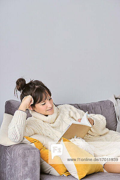 Frau liest ein Buch  während sie sich im Winter zu Hause auf dem Sofa ausruht
