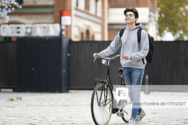 Smiling fashionable young man with backpack wheeling bicycle on footpath in city
