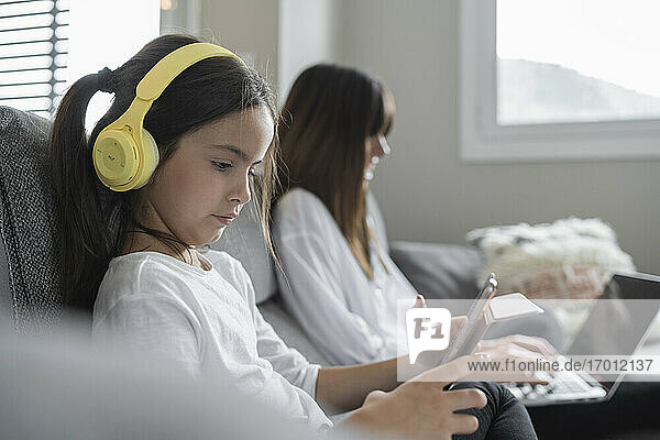 Tochter hält ein digitales Tablet  während die Mutter im Wohnzimmer sitzt und einen Laptop benutzt