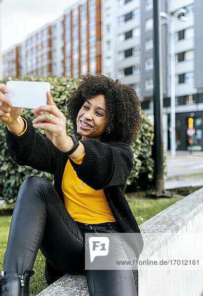 Lächelnde schöne Frau  die ein Selfie mit dem Mobiltelefon macht  während sie auf einem Geländer in der Stadt sitzt