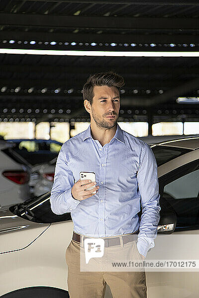 Männlicher Unternehmer mit Hand in der Tasche  der ein Mobiltelefon hält  während er auf einem Parkplatz steht
