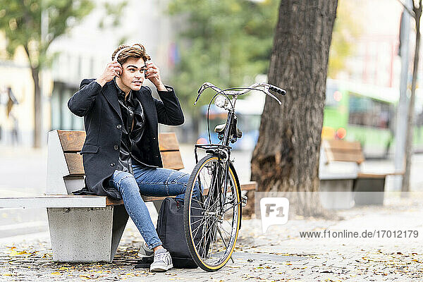 Junger Mann stellt drahtlose Kopfhörer ein  während er auf einer Bank mit dem Fahrrad in der Stadt im Herbst sitzt
