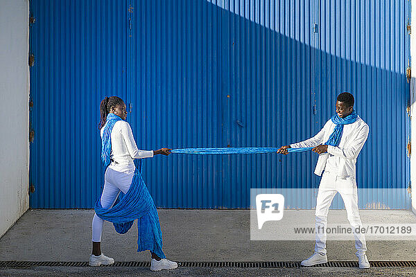 Heterosexual couple playing with blue fabric against corrugated wall on sunny day