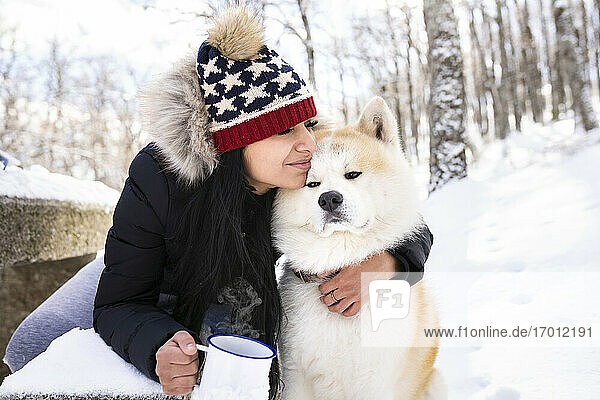 Frau mit geschlossenen Augen umarmt Akita-Hund und hält Kaffeetasse im Winter