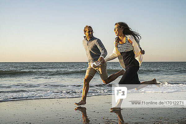 Fröhliches junges Paar  das sich beim Laufen am Strand während des Sonnenuntergangs an den Händen hält