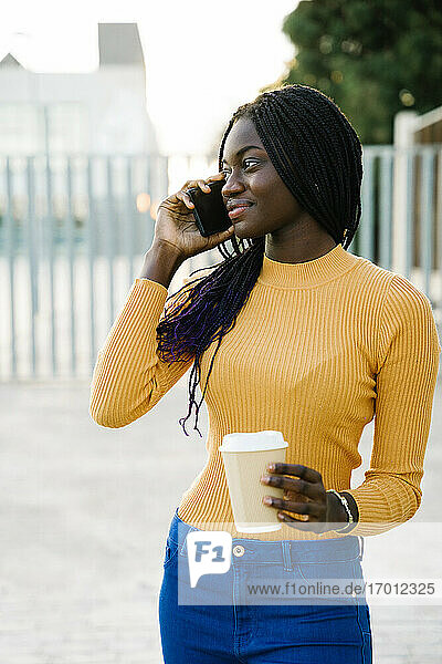 Smiling teenage girl with coffee cup looking away while talking on smart phone at street