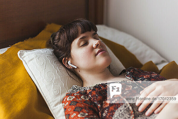 Junge Frau  die über drahtlose Kopfhörer eine geführte Meditation hört  während sie auf dem Bett liegt