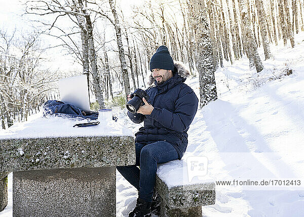 Smiling photographer using camera while sitting on rock bench in forest during snowy weather
