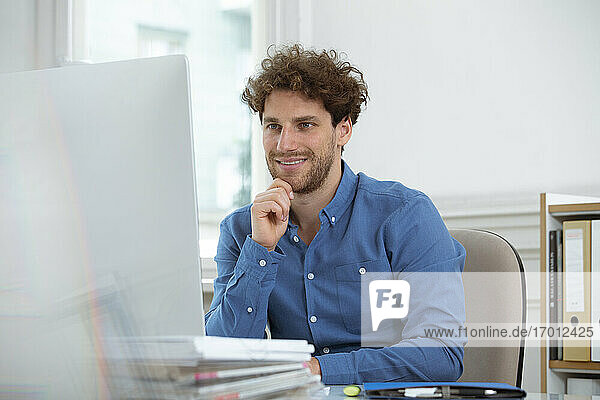 Lächelnder Geschäftsmann mit Hand am Kinn bei der Arbeit am Computer im Büro