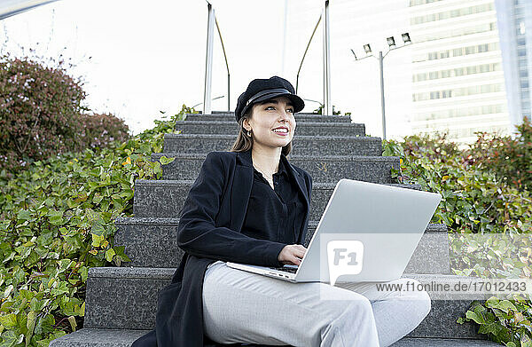 Junge Geschäftsfrau mit Mütze  die wegschaut  während sie mit einem Laptop auf einer Treppe sitzt
