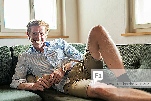 Lächelnder Mann mit Kaffeetasse auf der Couch im Wohnzimmer liegend