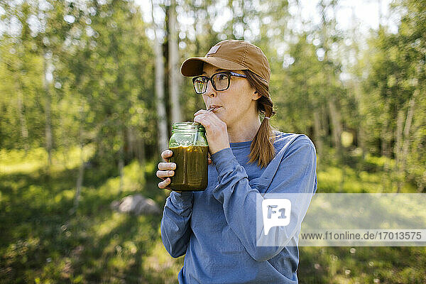USA  Utah  Uinta National Park  Woman wearing eyeglasses and baseball cap drinking coffee from jar in forest