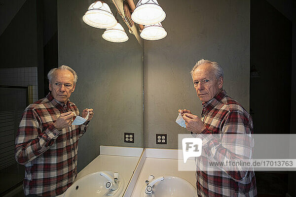Älterer Mann hält Covid-Schutzmaske vor dem Spiegel im Badezimmer