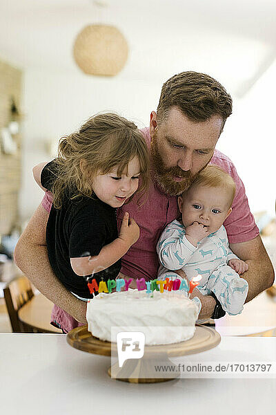 Vater trägt Kinder (2-3  6-11 Monate) und pustet Geburtstagskerzen auf Kuchen