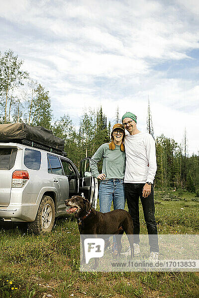 USA  Utah  Uinta National Park  Portrait of smiling couple with dog standing in meadow  off road car in background