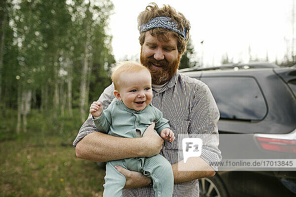 USA  Utah  Uinta National Park  Lächelnder Mann hält Baby-Sohn (6-11 Monate) im Feld  Auto im Hintergrund