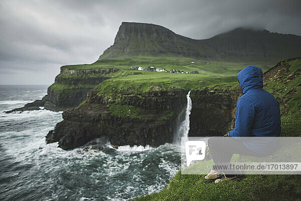 Dänemark  Färöer Inseln  Gasadalur Dorf  M-Lafossur Wasserfall  Mann sitzt am Rande der Klippe und schaut auf Mulafossur Wasserfall