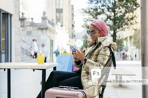 Lächelnde Frau mit rosa Haaren  die ein Mobiltelefon benutzt  während sie in einem Straßencafé sitzt