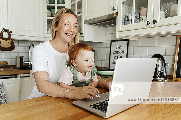 Frau mit kleiner Tochter benutzt Laptop am Küchentisch