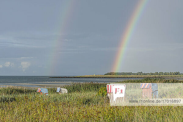 Doppelter Regenbogen  der sich über Strandkörbe mit Kapuze wölbt  die am grasbewachsenen Ufer stehen