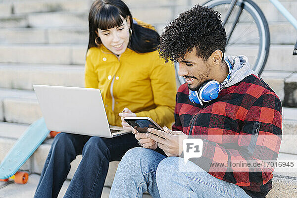 Junger Mann  der ein digitales Tablet benutzt  während er neben einer Freundin sitzt  die einen Laptop auf einer Treppe benutzt