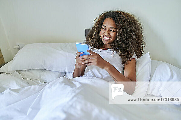 Glückliche junge Frau  die ihr Smartphone benutzt  während sie sich auf dem Bett im Schlafzimmer zu Hause entspannt