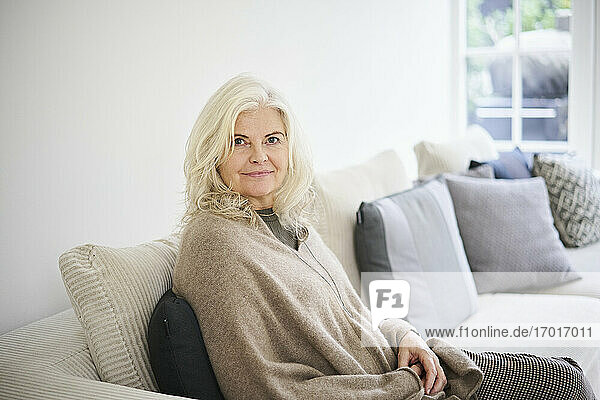 Schöne ältere Frau mit langen weißen Haaren sitzt auf Sofa im Wohnzimmer zu Hause