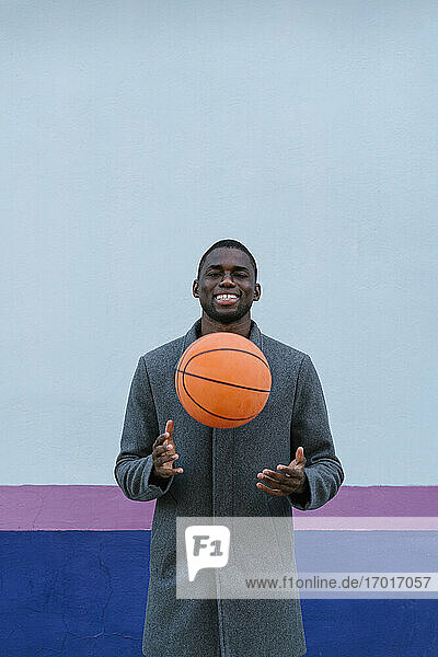 Junger Mann lächelnd beim Spielen mit Basketball gegen mehrfarbige Wand