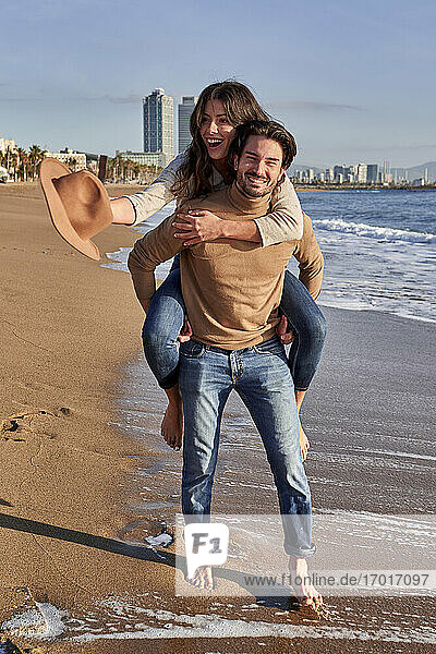 Lächelnder Mann  der eine sorglose Frau huckepack nimmt  während er am Strand steht