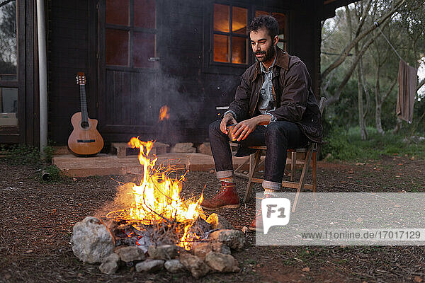 Junger Mann mit Kaffeetasse sitzt auf einem Stuhl in der Nähe eines Lagerfeuers bei einem Holzhaus