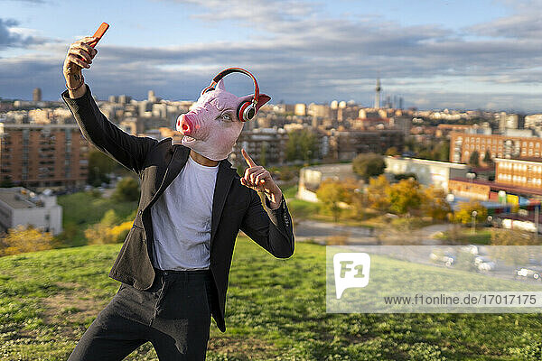 Geschäftsmann trägt Schweinemaske und drahtlose Kopfhörer  während er ein Selfie mit seinem Smartphone auf einem Hügel macht