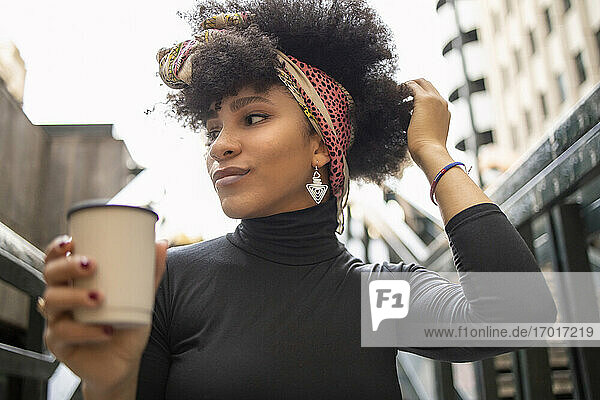 Afro young woman with hand in hair looking away while holding disposable coffee cup in city