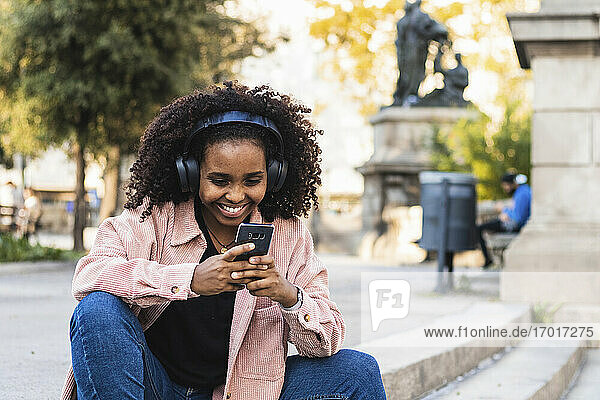 Happy afro young woman using mobile phone while listening music on staircase in city