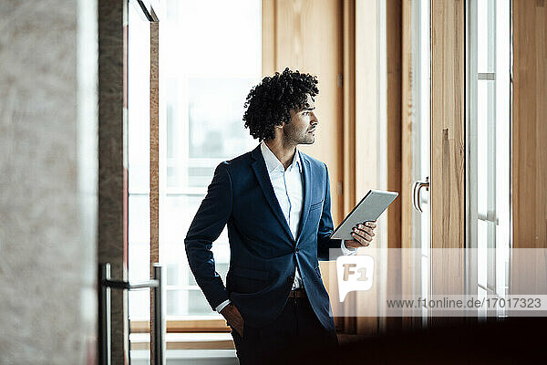 Nachdenklicher männlicher Unternehmer  der ein digitales Tablet in der Hand hält  während er am Arbeitsplatz wegschaut