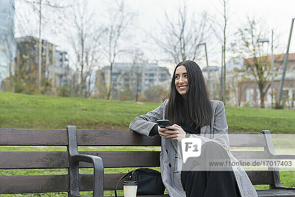 Lächelnde weibliche Fachkraft mit Kaffeetasse  die wegschaut  während sie auf einer Parkbank ihr Handy hält
