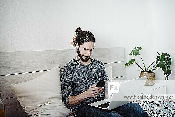 Junger Mann telefoniert  während er mit seinem Laptop zu Hause auf dem Bett sitzt