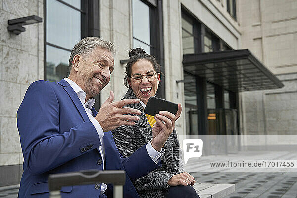 Cheerful male and female entrepreneurs sharing smart phone while sitting against building