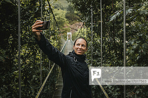 Lächelnde Frau  die ein Selfie mit ihrem Smartphone macht  während sie auf einer Hängebrücke im Wald steht