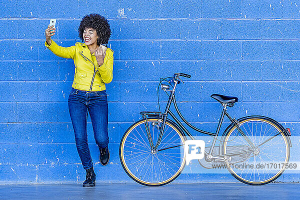 Fröhliche junge Frau mit Einwegbecher nimmt Selfie gegen blaue Wand