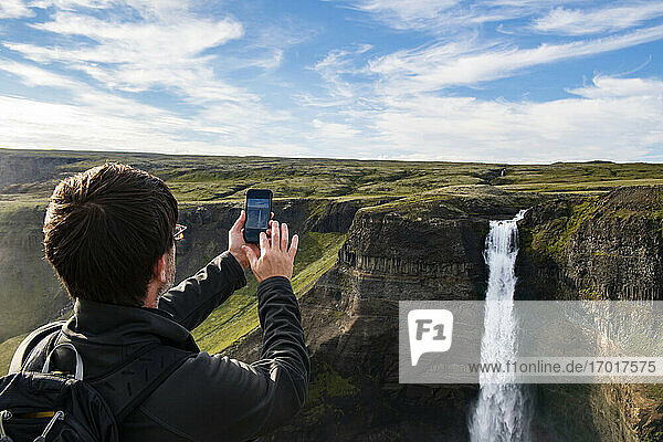 Tourist fotografiert den Haifoss-Wasserfall mit dem Mobiltelefon  während er in Island steht