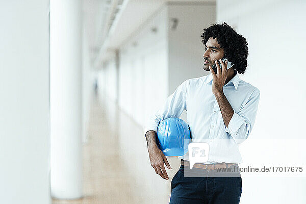 Junger männlicher Architekt mit Schutzhelm  der wegschaut  während er im Büro auf dem Flur mit seinem Handy telefoniert