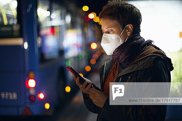 Frau mit Gesichtsmaske  die an einer Bushaltestelle steht und ein Mobiltelefon benutzt