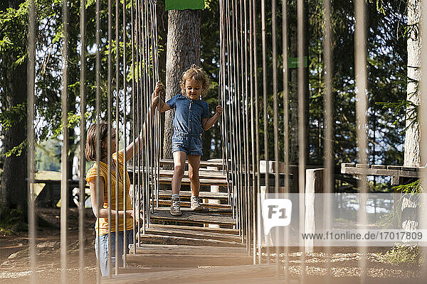 Mother assisting little daughter walking across small suspension bridge on forest obstacle course
