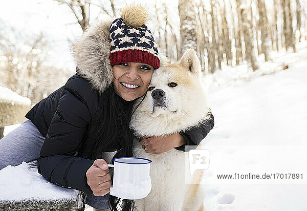 Glückliche Frau mit Kaffeetasse  die ihren Hund im Schnee umarmt