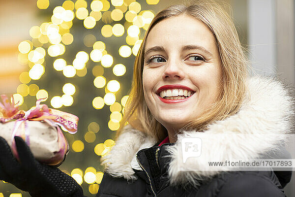 Lächelnde Frau mit blondem Haar  die ein Geschenk gegen die Weihnachtsbeleuchtung hält
