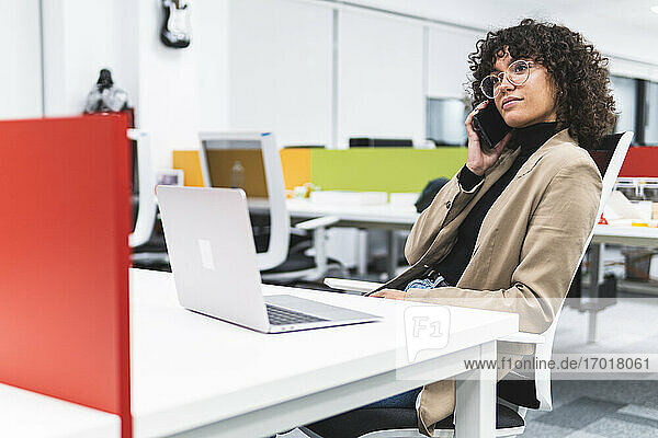 Female entrepreneur with laptop on desk talking on mobile phone at work place