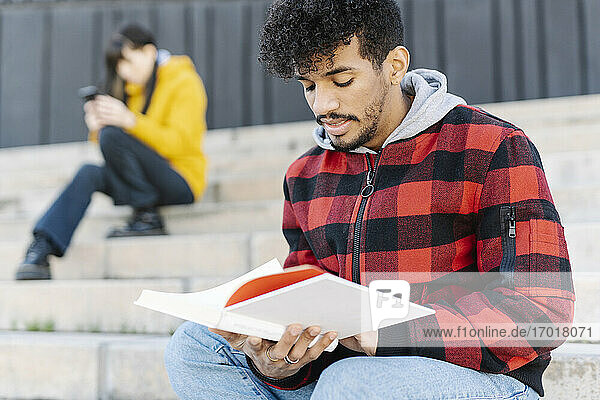 Junger Mann mit Kapuzenshirt liest ein Buch  während er auf einer Treppe neben einem Skateboard sitzt