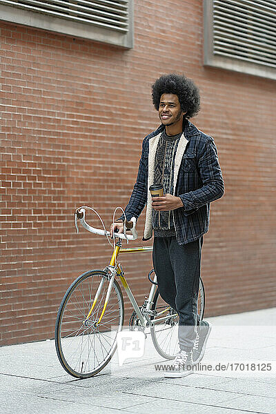 Lächelnder junger Mann mit Kaffee in der Hand  während er mit dem Fahrrad auf der Straße an einer Backsteinmauer spazieren geht