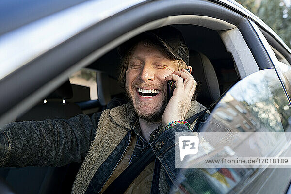 Nahaufnahme eines fröhlichen Mannes  der im Auto über sein Smartphone spricht  gesehen durch ein Fenster